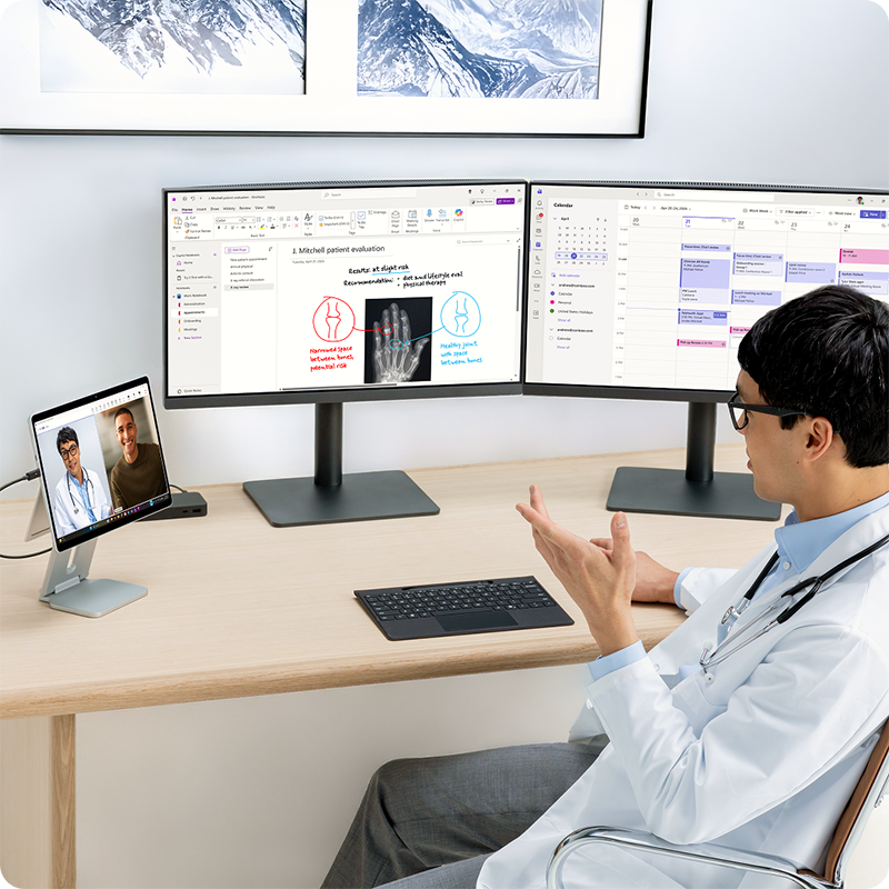 一名医生坐在办公桌前进行视频通话，使用 Surface Pro 商用版的摄像头，同时在多台显示器上开展工作。